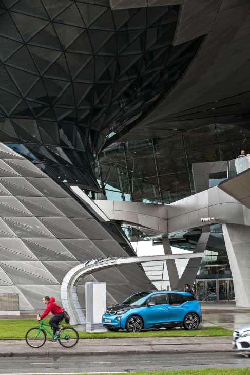 Blue BMW i3 electric vehicle parked in front of modern architecture with cyclist riding past.