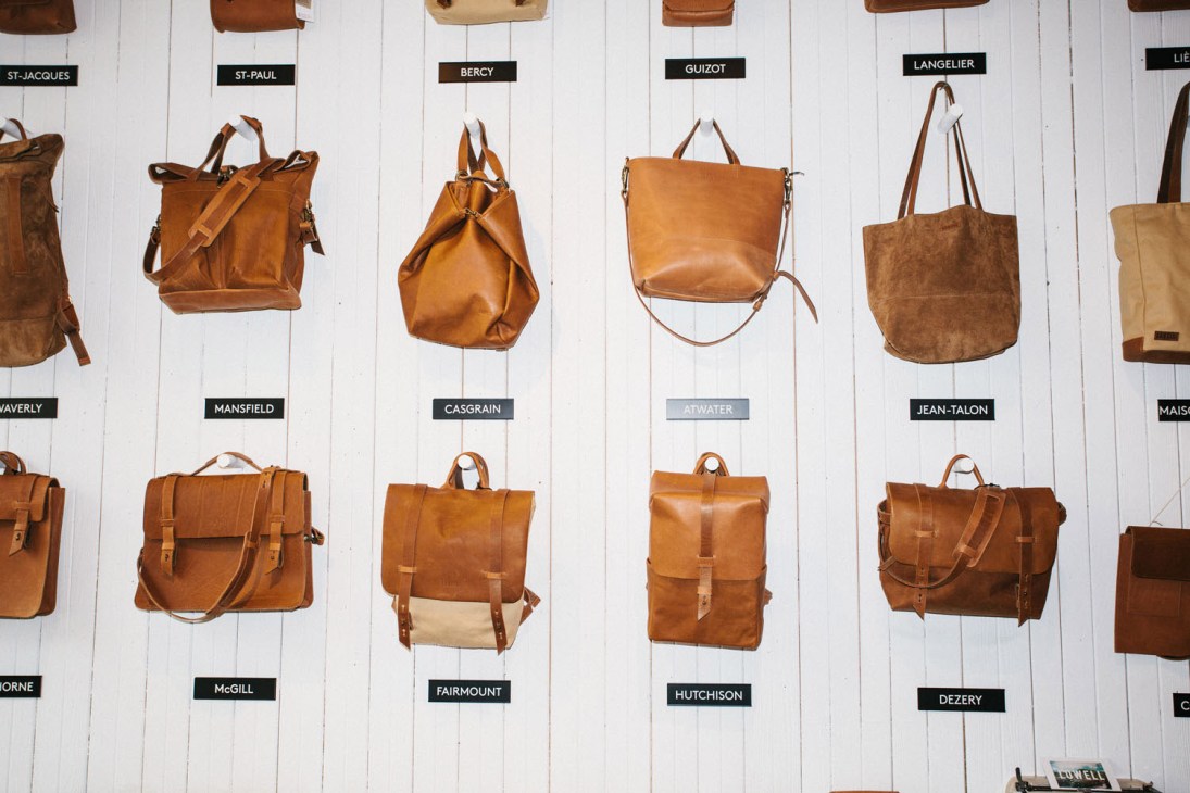 Leather bags and backpacks displayed on white wall with neighborhood labels at Lowell in Montreal's Mile End.