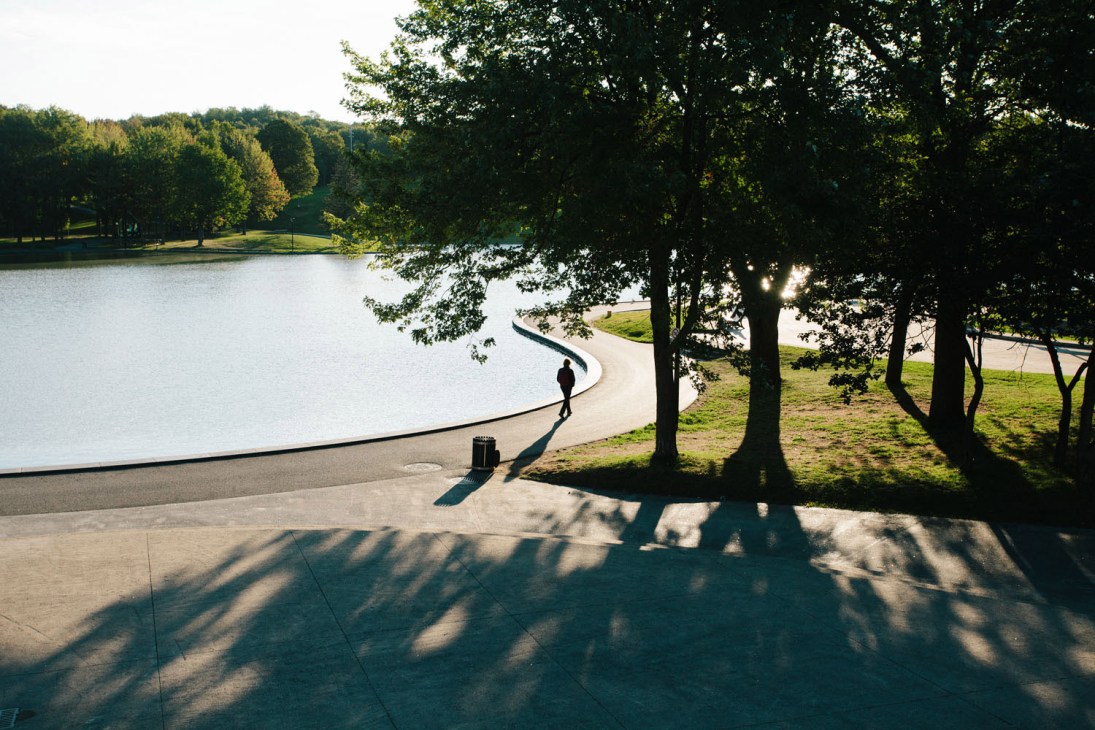Person jogging along a curved waterside path surrounded by mature trees and green lawns.
