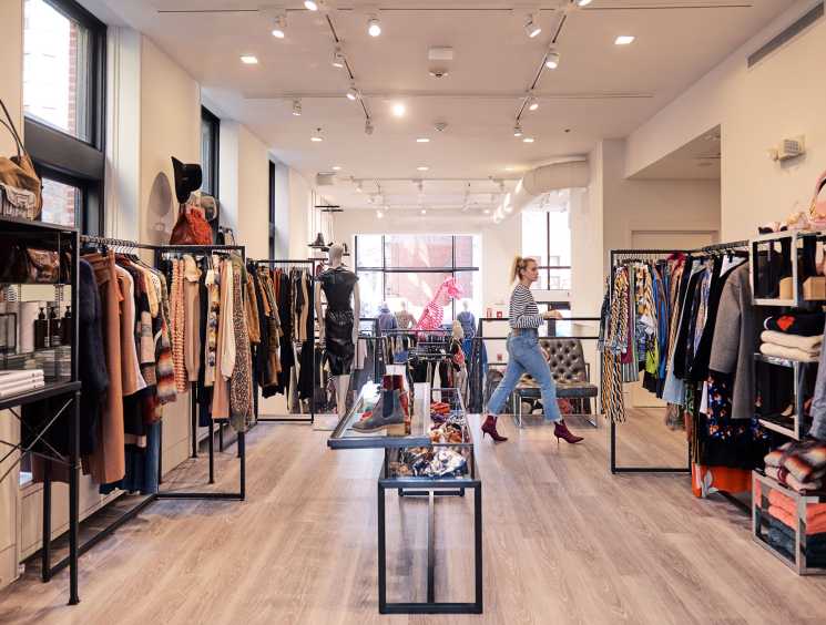 Interior of All Too Human boutique showing clothing racks, designer merchandise, and a female shopper browsing in a minimalist retail space.