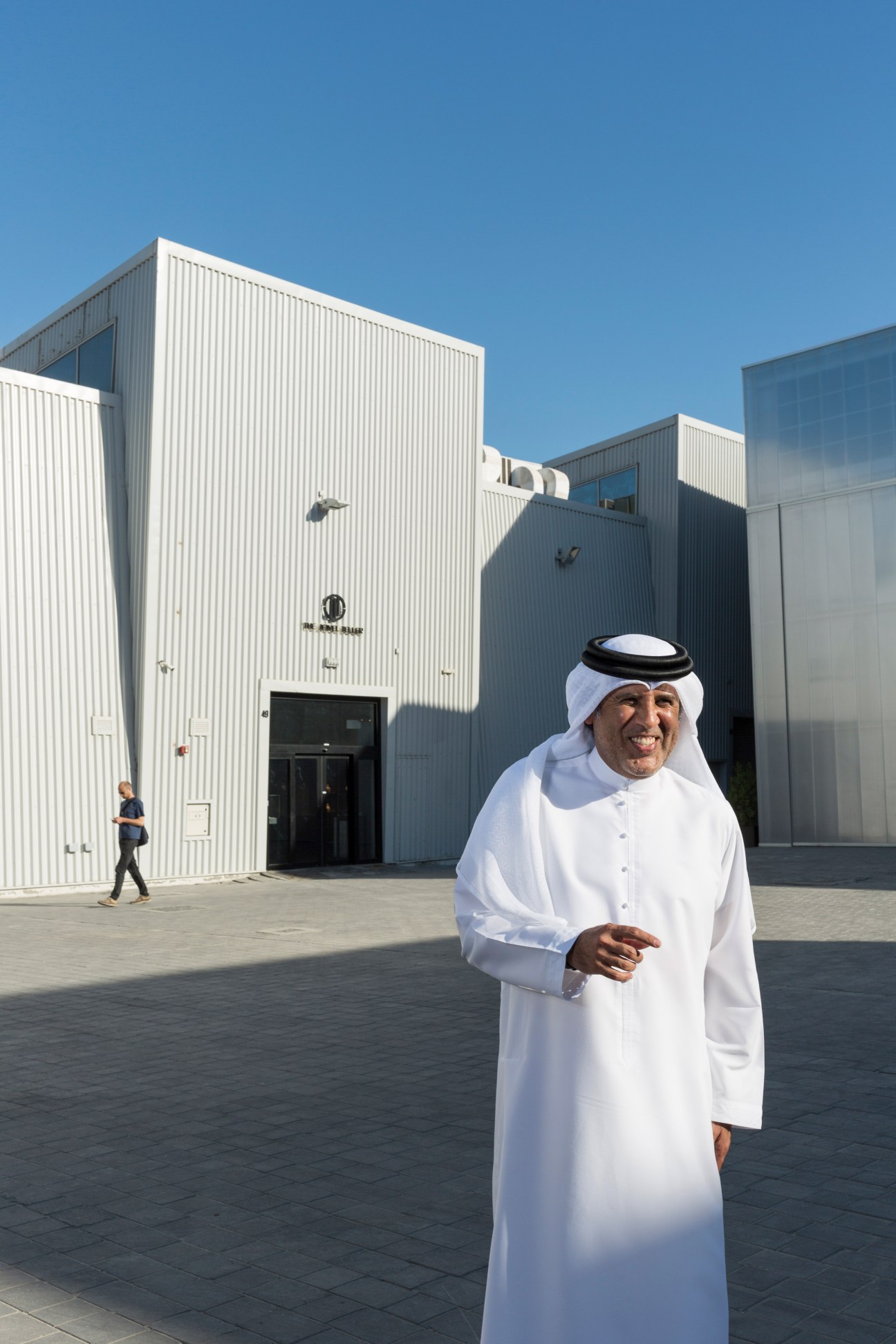 Man in white traditional Gulf dress standing in front of modern industrial warehouse buildings.