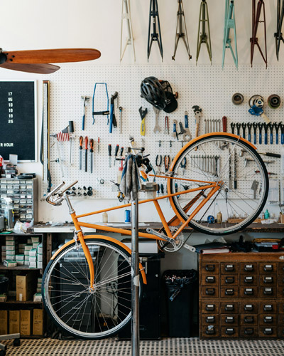 Orange vintage-inspired bicycle displayed in Heritage Bicycles General Store with tools and supplies on white pegboard wall.