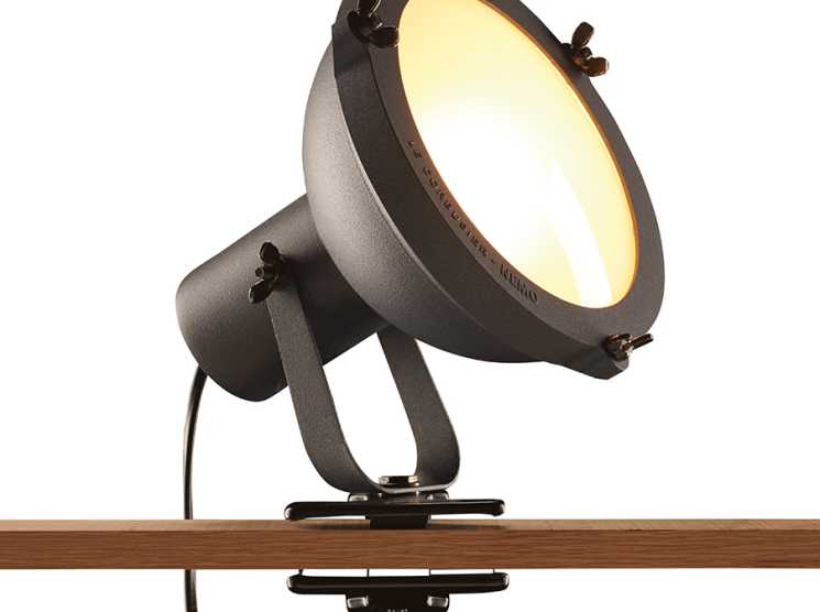 Nemo clip lamp with adjustable dark metal head and glowing warm light mounted on wooden shelf edge.