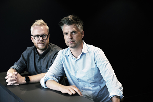 Two men posing for a portrait against a dark background; George Kay on the right wearing a light blue shirt and Jim Field Smith on the left wearing a dark shirt.