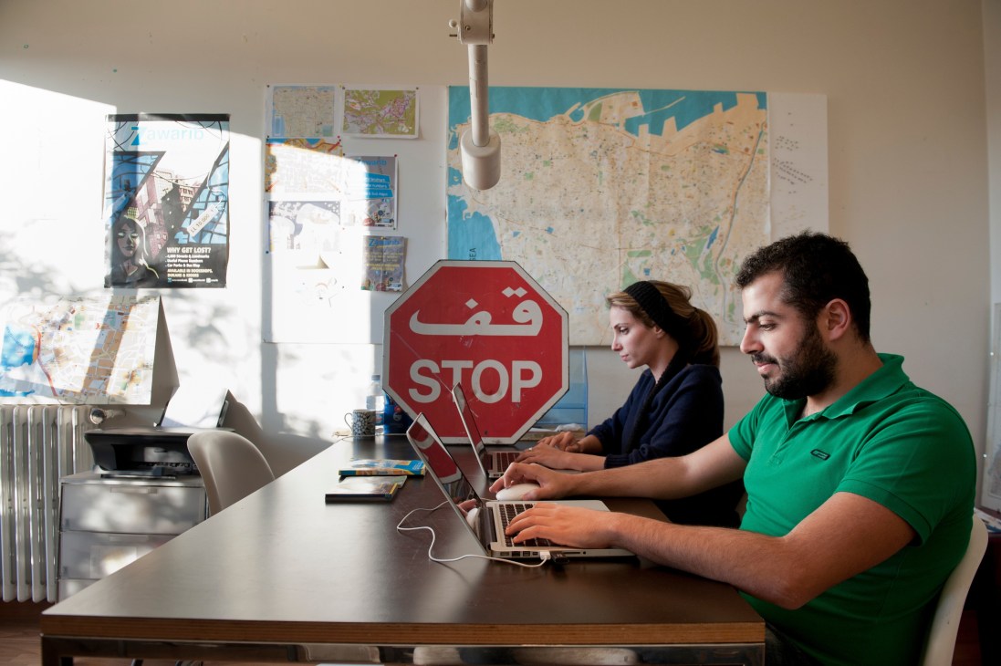 Two people work at laptops in an office with maps and a bilingual stop sign on the wall.