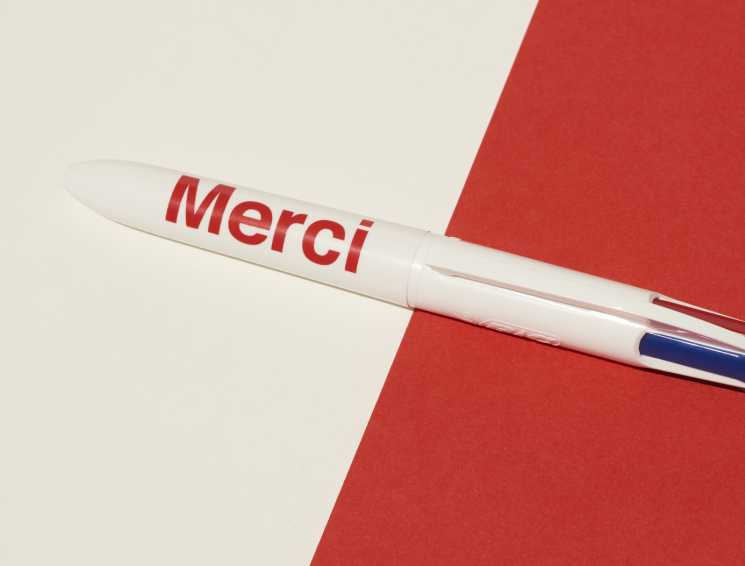 Merci retractable pen with white barrel and red text on red and cream background.