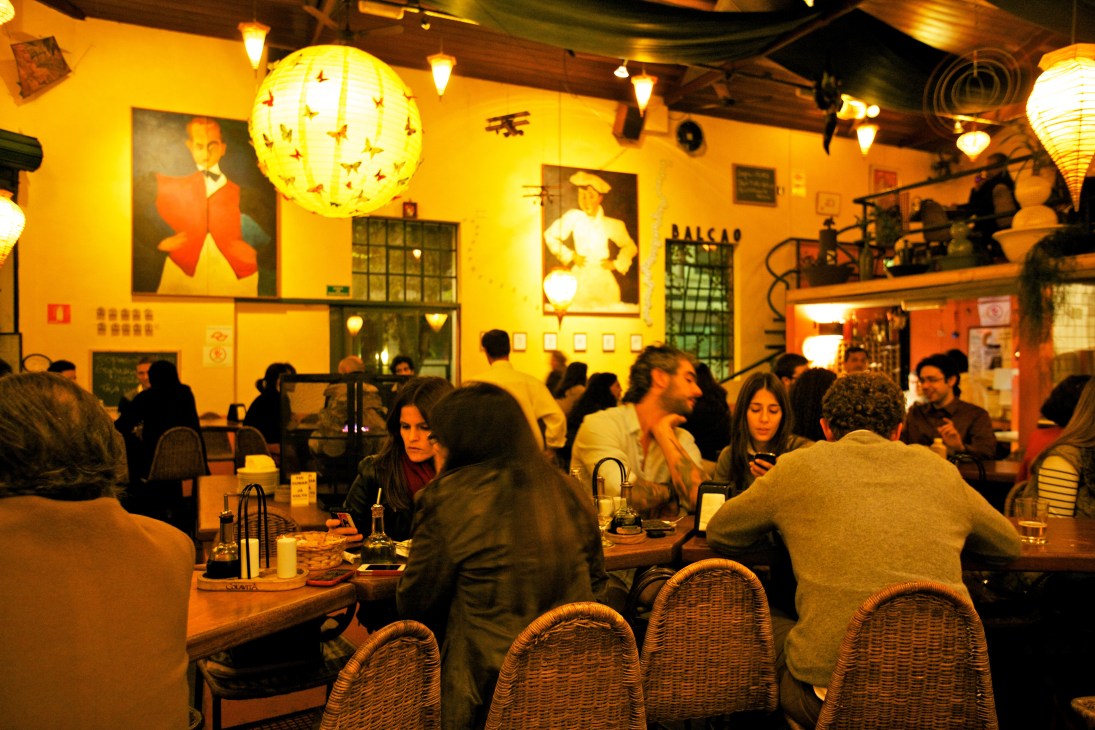 Interior of Bar Balcão in São Paulo with diners at communal counter seating under warm yellow lighting and artwork.