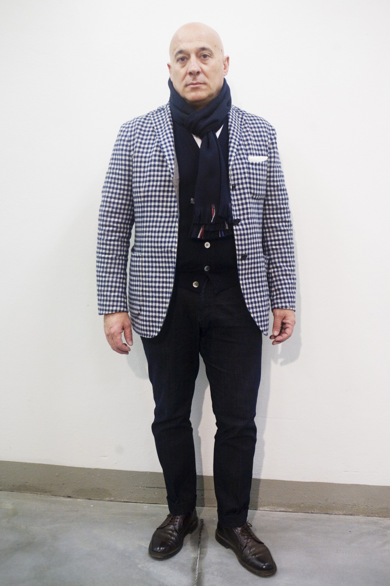 Man wearing blue gingham blazer, navy scarf, black trousers and dark shoes standing against white wall.
