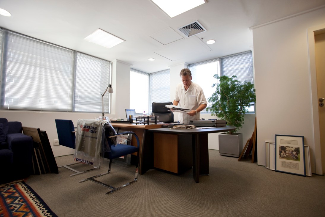 Man working at desk in bright office with windows, chair, and potted plant.