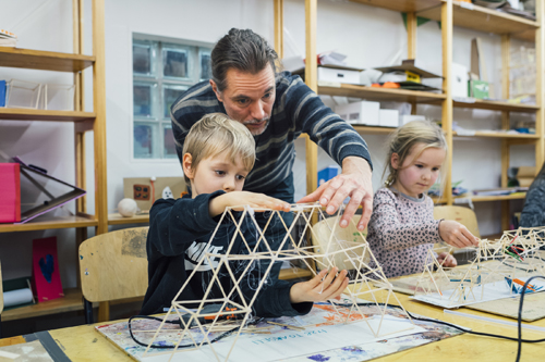 An adult and two children building a wooden structure together at a table with craft materials.