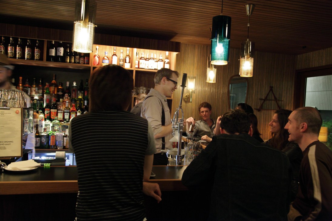 Bartender serves customers at Fuglen bar in Tokyo with wooden interior and pendant lighting.