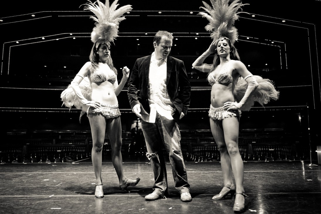 Black and white photograph of a male performer in a blazer dancing with two female showgirls in elaborate feathered headdresses and sequined costumes on a lit stage.