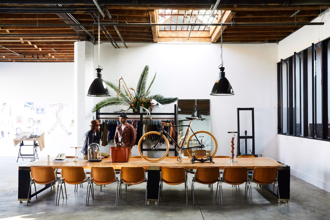 The Space in Brooklyn features a long wooden table with tan leather chairs, bicycles, clothing racks, and hanging black pendant lights in an industrial loft setting.