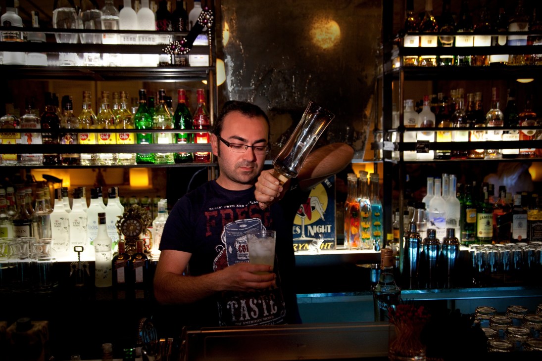 Bartender Ahmet Boyaci mixes a cocktail behind the bar at Baylo in Istanbul, with shelves of spirits visible behind him.