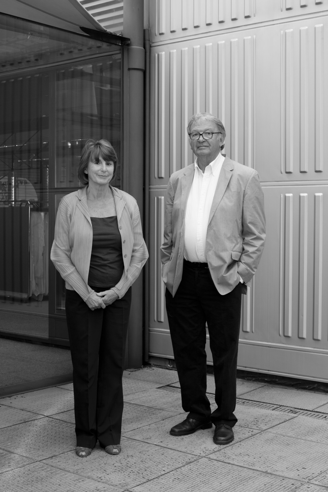 Two people standing in front of a modern building with white paneled doors and windows.