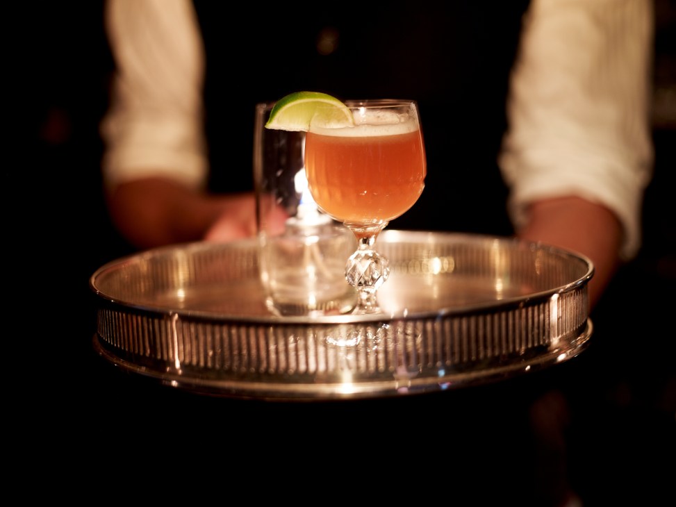 Bartender presenting an amber cocktail with lime garnish on a brass-topped table.