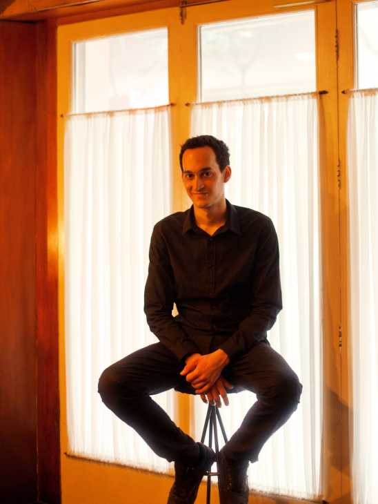Man in black shirt sitting on stool against orange-framed windows with white curtains.