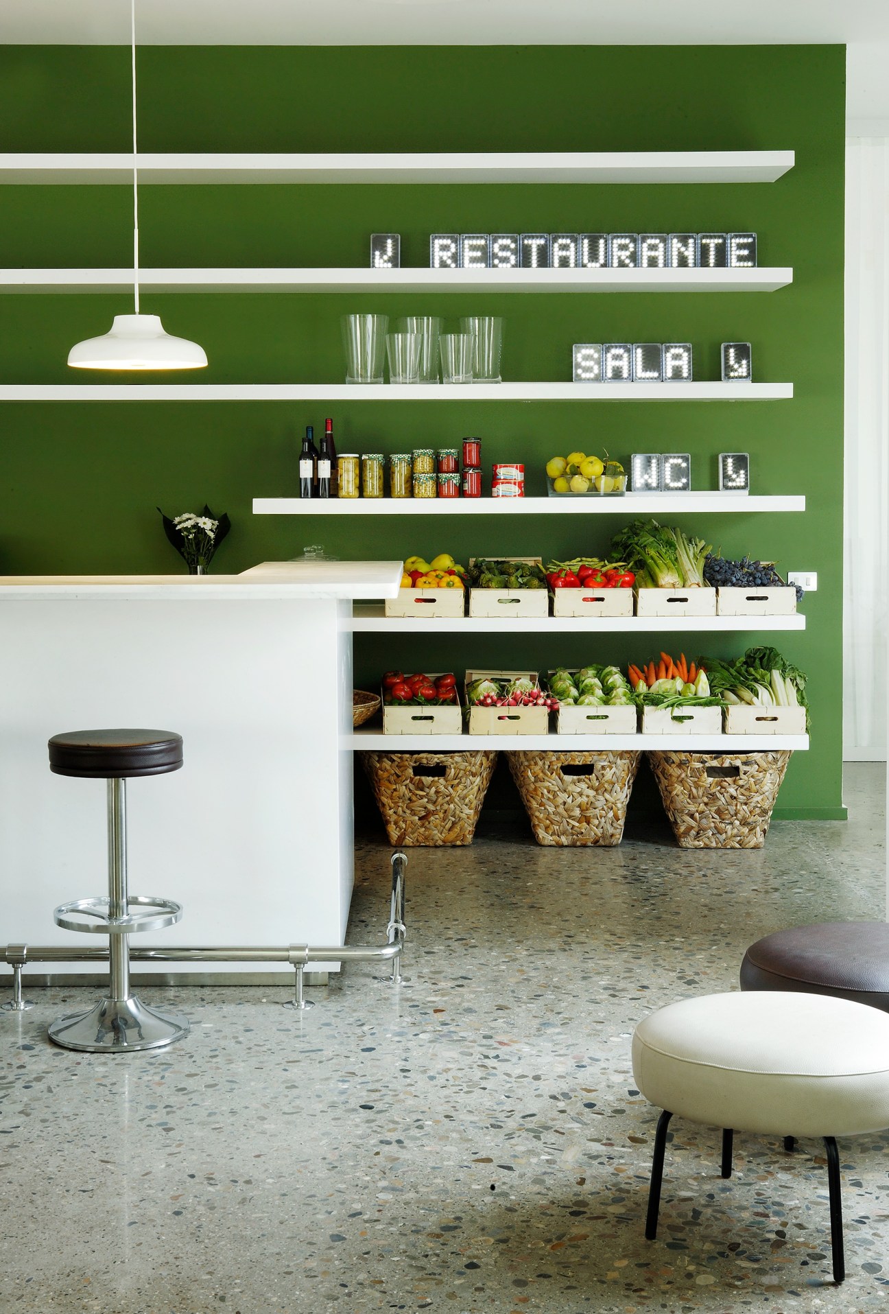 Modern restaurant interior with green wall, white shelving, fresh produce displays and minimalist seating.