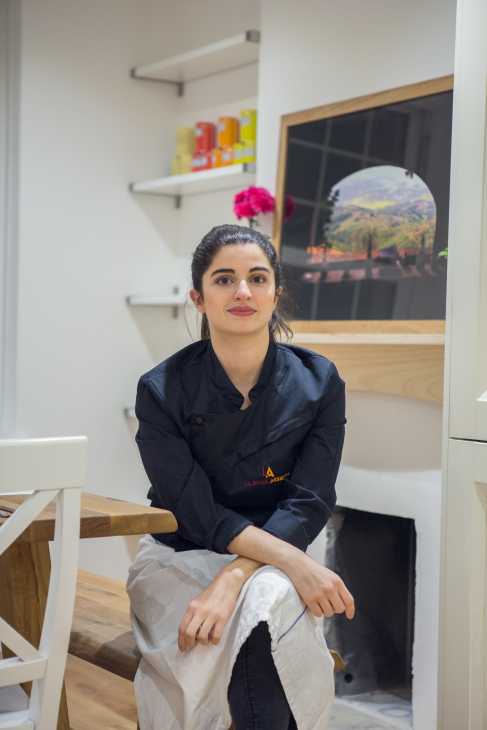 Woman in La Belle Assiette chef's jacket sits in modern kitchen with white shelving and artwork.