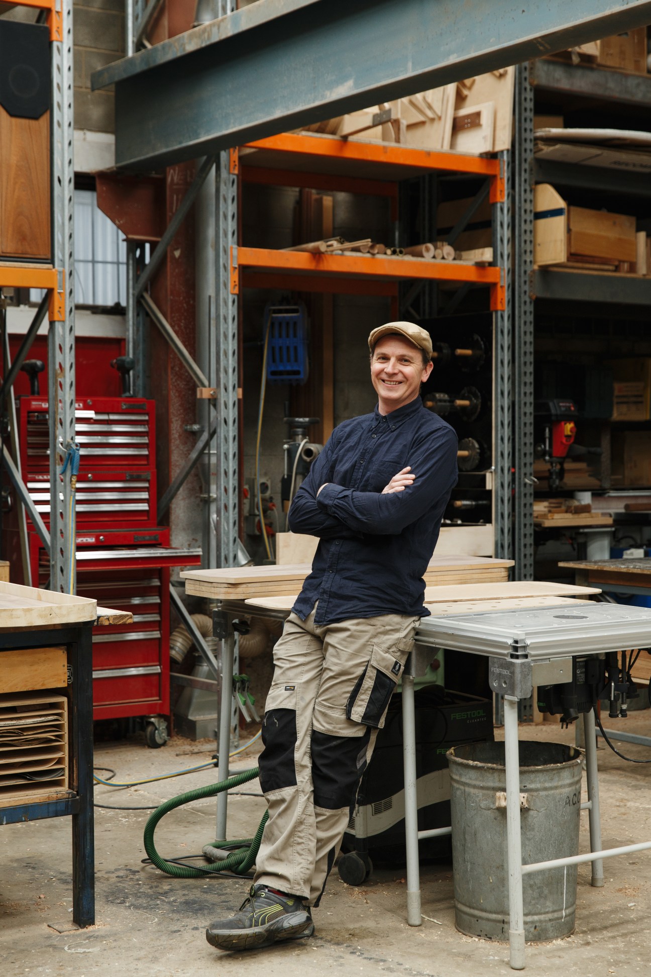 Man in cap and navy shirt standing with crossed arms in woodworking workshop with shelving and tools