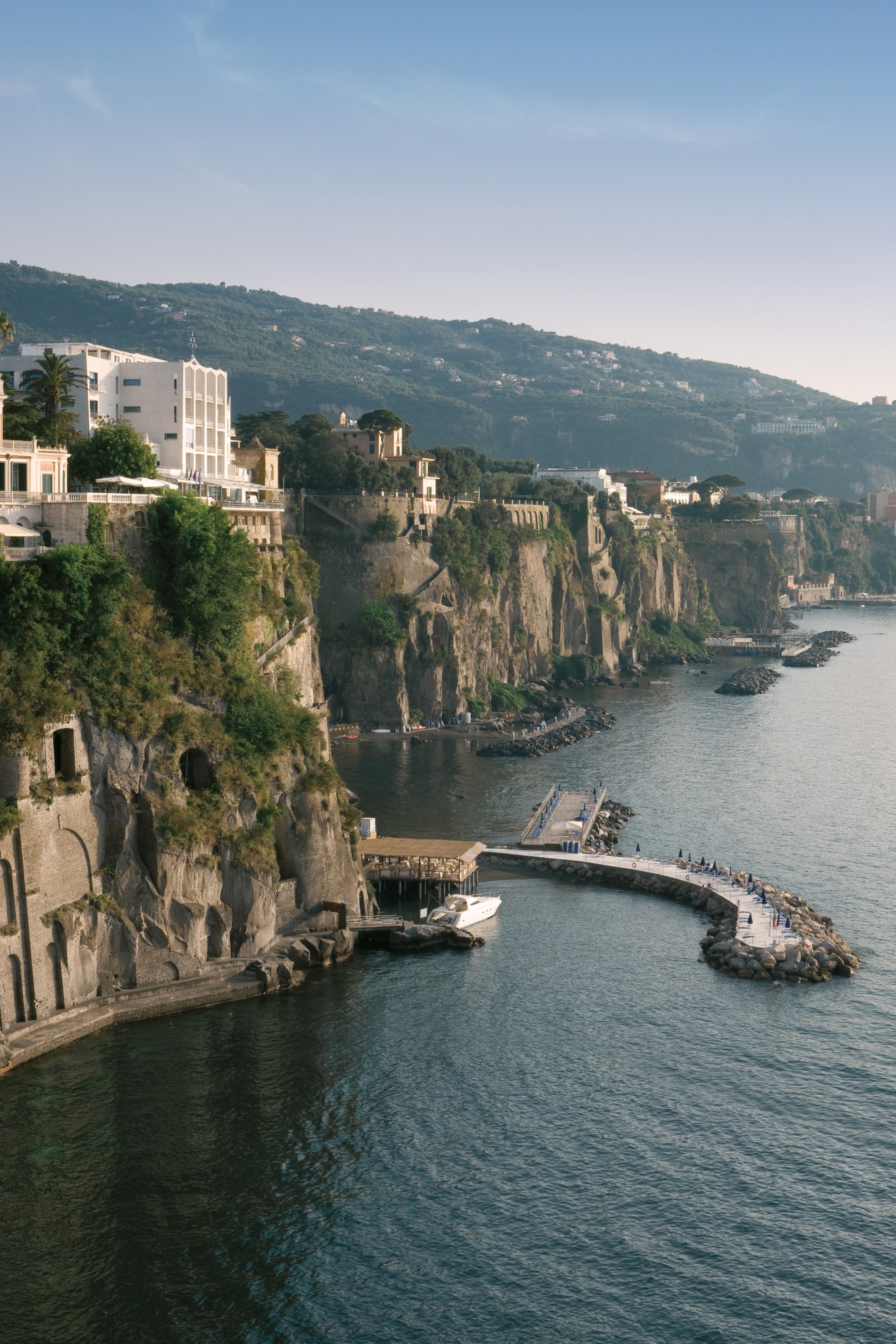 Clifftop hotel with white modernist building overlooking Mediterranean Sea and coastal town nestled on rocky peninsula.
