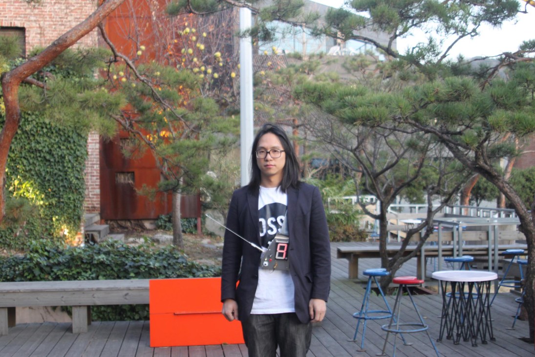 Young man with glasses wearing black blazer standing in outdoor courtyard with brick building and trees.