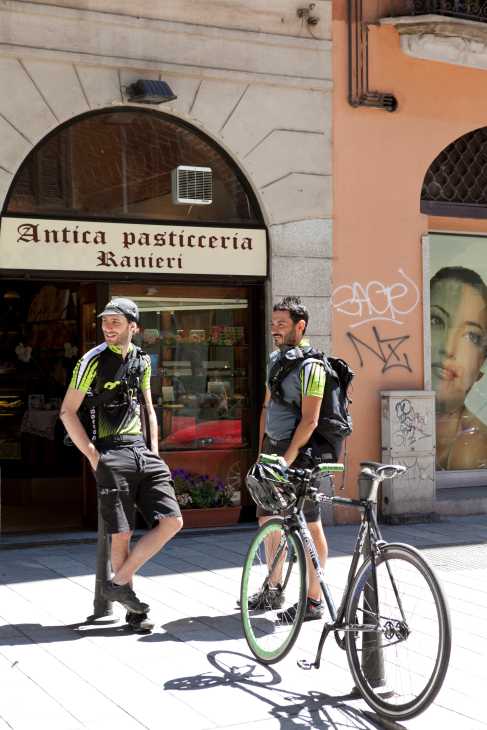 Two UBM cyclists in neon yellow and black gear stand with their bikes outside a Milan pastry shop.