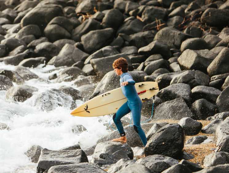 A surfer in a blue wetsuit carrying a surfboard walks across rocky coastline with crashing waves