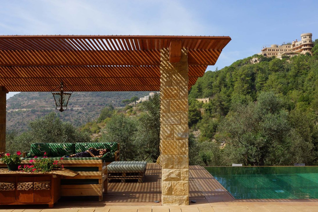 Guest relaxing under a wooden pergola by a pool with Mediterranean views and castle ruins visible on hillside.