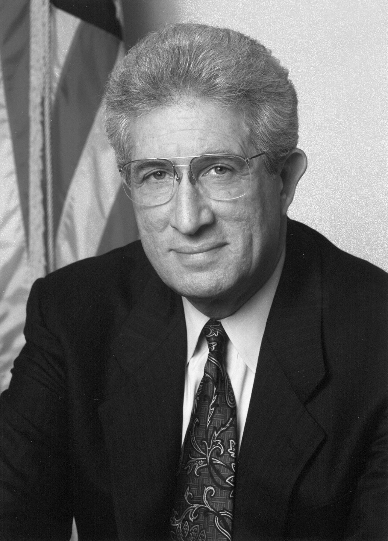 Black and white portrait of Richard H Solomon, an older man wearing glasses, dark suit and patterned tie.