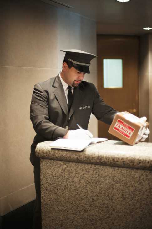 Joe Toro, doorman at 45 East 80th Street in New York, wearing uniform and white gloves while handling a package at the building's desk.
