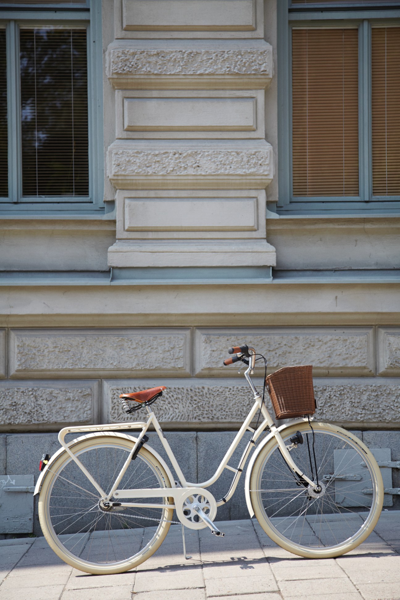 Cream-coloured Pilen Special bicycle with brown leather saddle and wicker basket parked against a stone building facade.