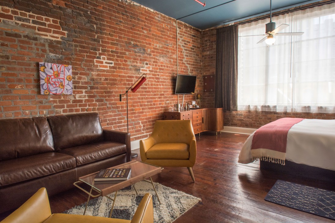 Modern hotel suite with exposed brick walls, brown leather sofa, mustard armchair, and bed with pink throw.