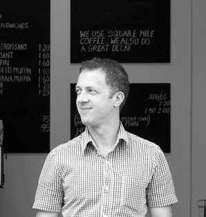 Ben Townsend, owner and barista at The Espresso Room in London, smiling in front of a coffee shop menu board.