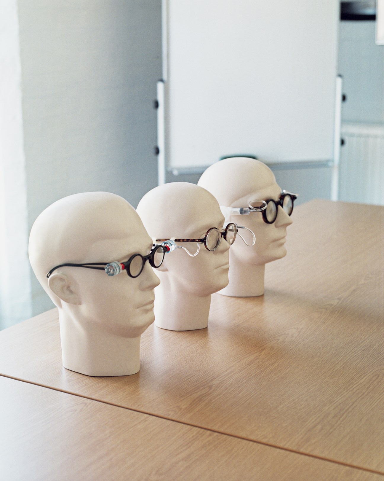 Three mannequin heads displaying adjustable eyeglasses with dials on wooden table.