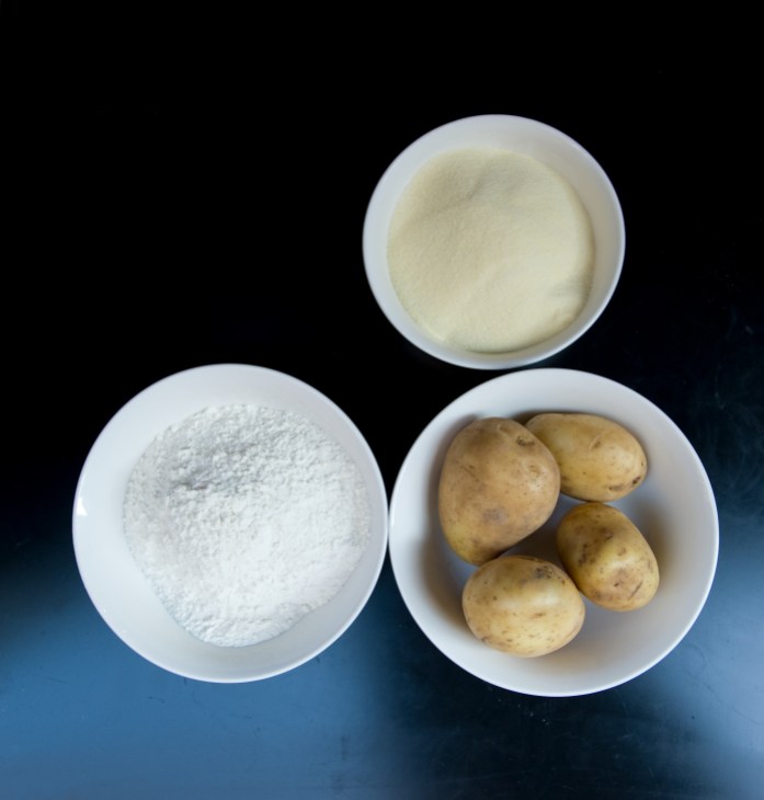 Three white bowls containing ingredients for homemade potato gnocchi: flour, mashed potatoes, and whole potatoes.