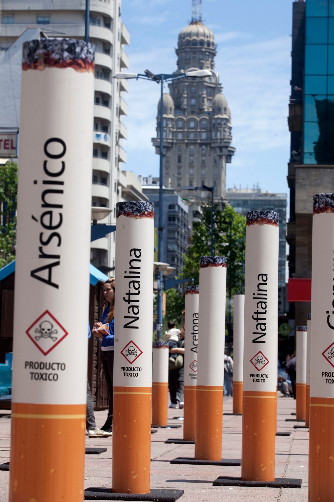 Giant cigarette sculptures with toxic product warnings displayed in an urban plaza with historic architecture.