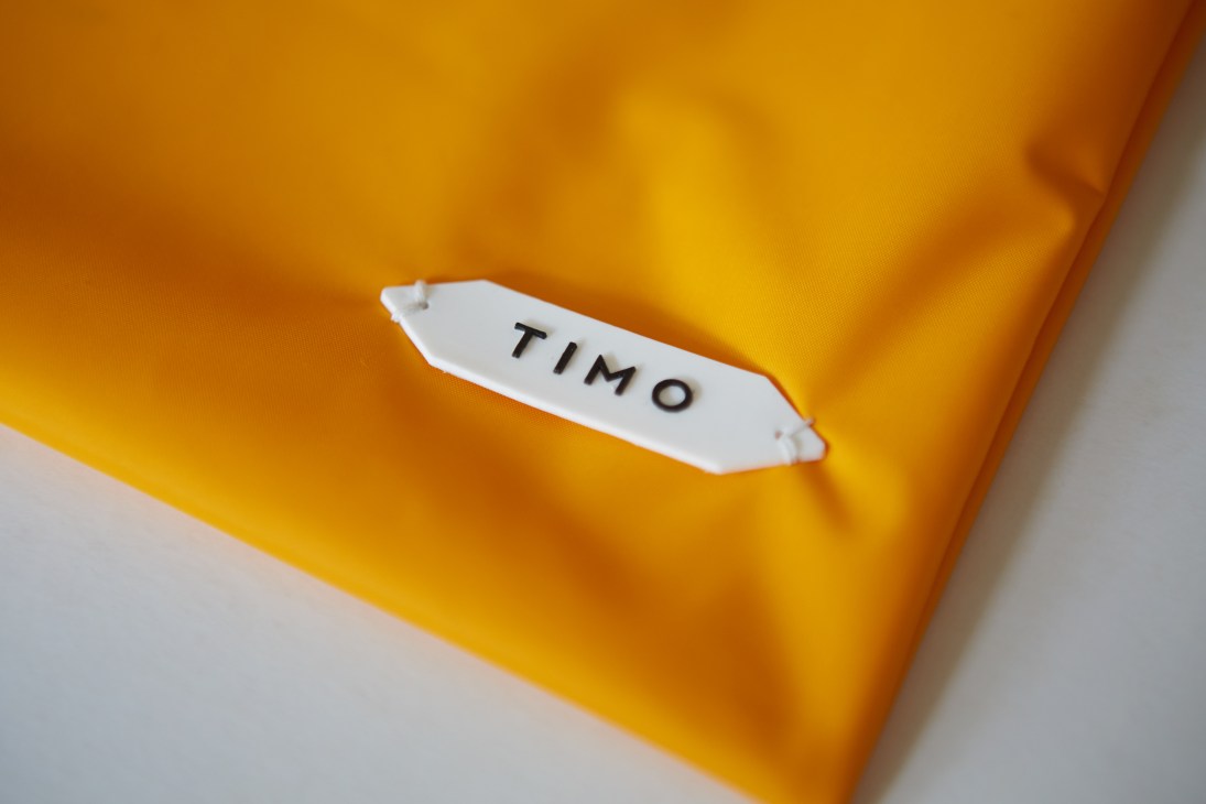 White fabric label reading 'TIMO' sewn onto bright yellow-orange swim trunk material.