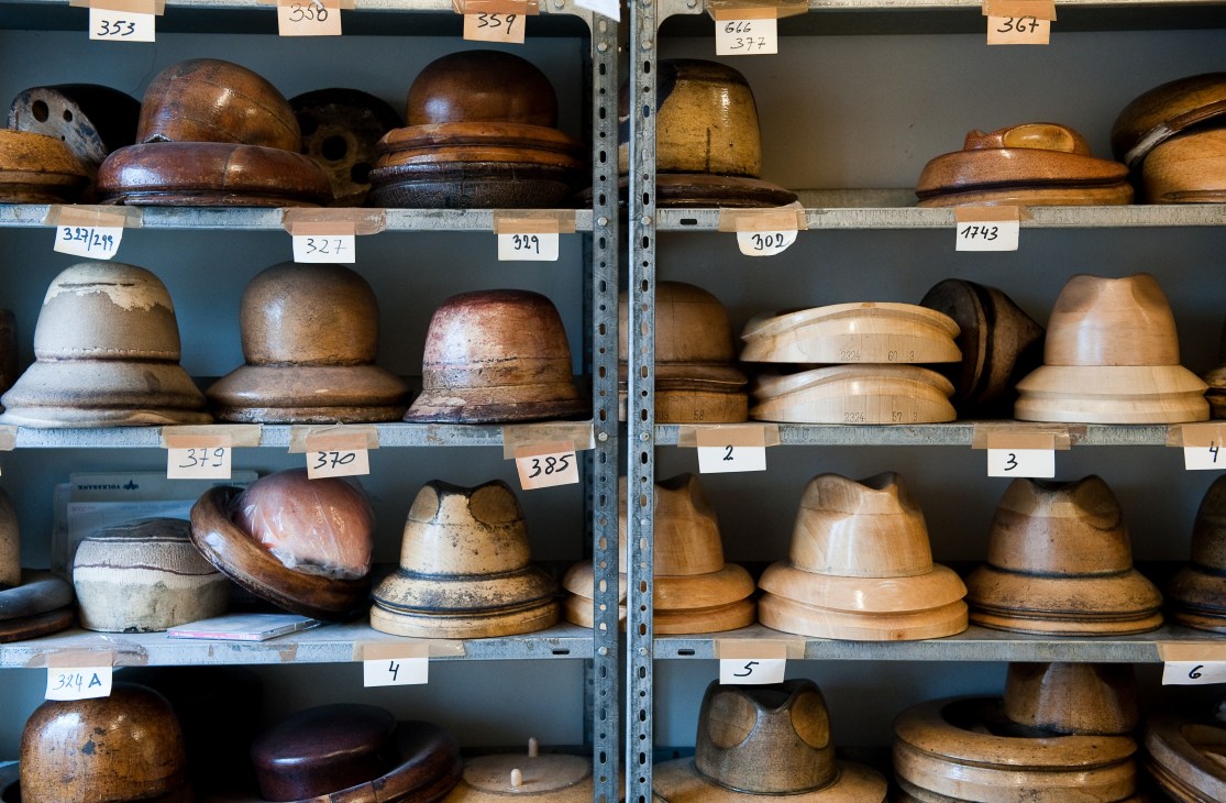 Wooden hat moulds of various sizes arranged on metal shelves with numbered labels at Mühlbauer atelier in Vienna.