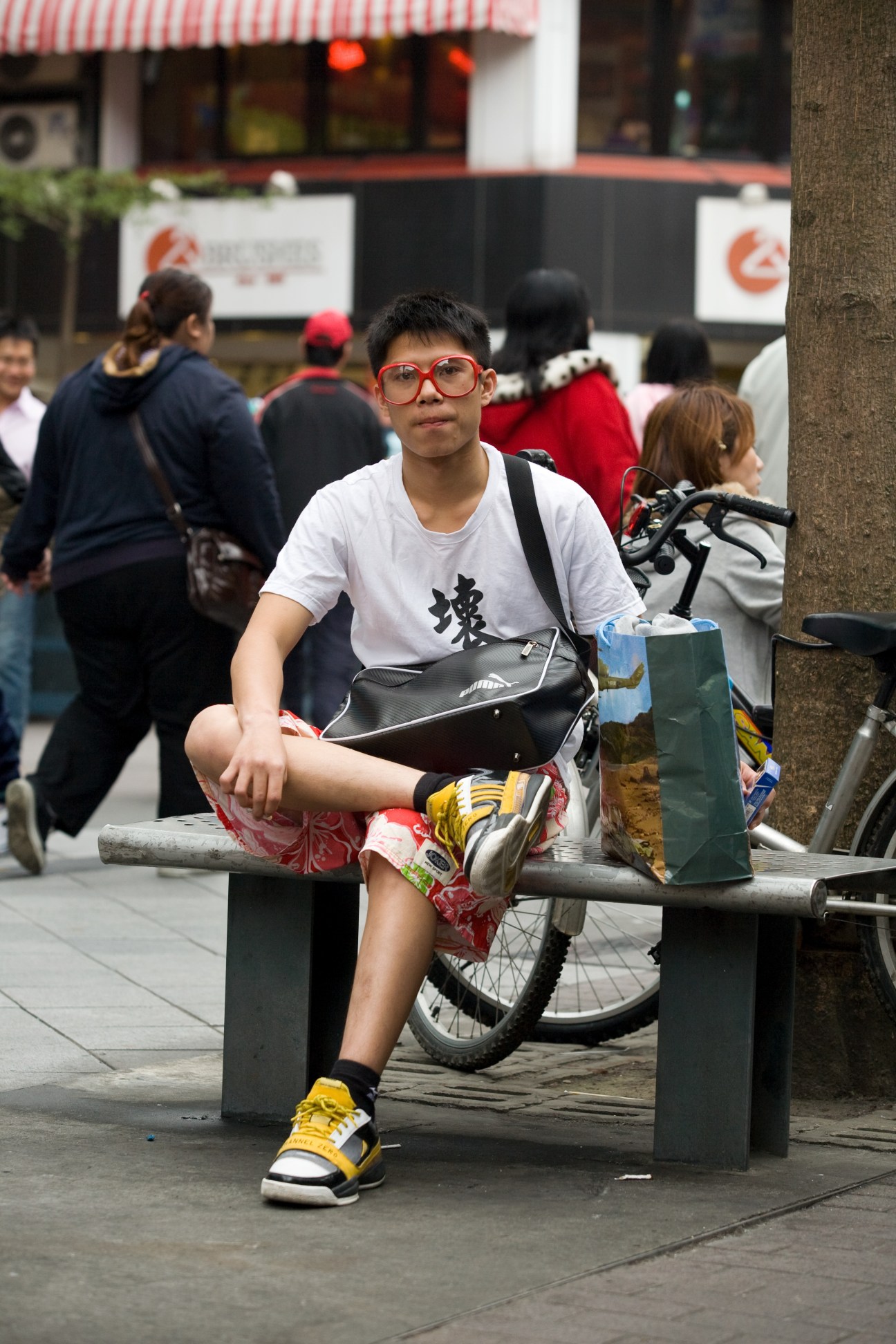 Young person wearing red-framed glasses and white t-shirt sitting on bench in Taipei street with bike and crowd behind