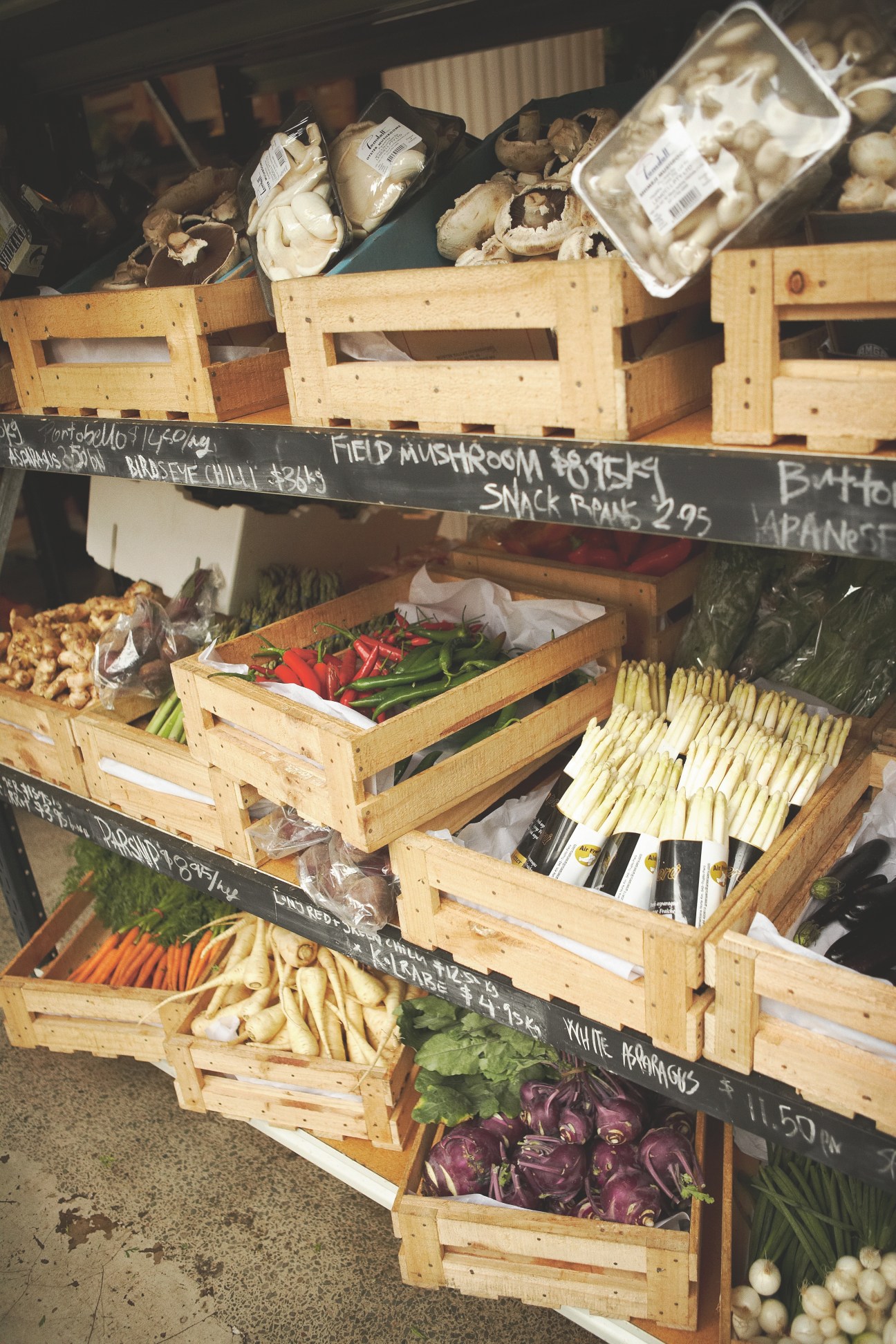 Wooden produce crates with packaged mushrooms and fresh vegetables displayed on tiered shelving with handwritten price signs.