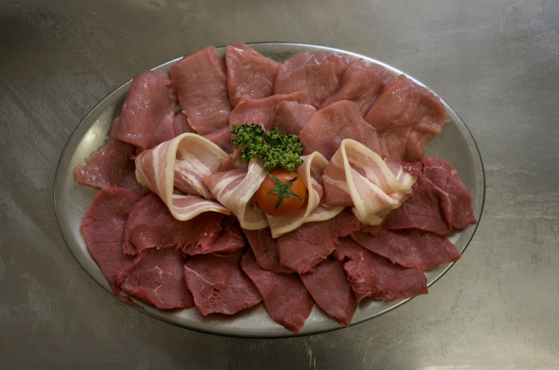 Thinly sliced raw Simmental beef, veal and bacon arranged on a plate with parsley, tomato and bacon garnish.