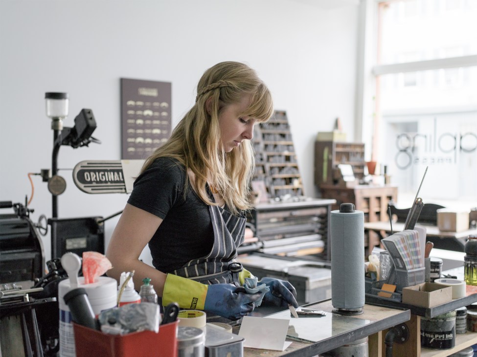 Woman working at letterpress printing machine in Sapling Press studio with architectural models in background.