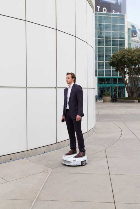 Man in dark suit standing on Ford's Carr-E rideable platform in an urban plaza.