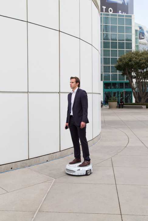 Man in dark suit standing on Ford's Carr-E rideable platform in an urban plaza.