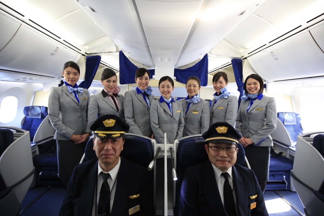 ANA flight crew and staff pose in aircraft cabin wearing gray and blue uniforms.