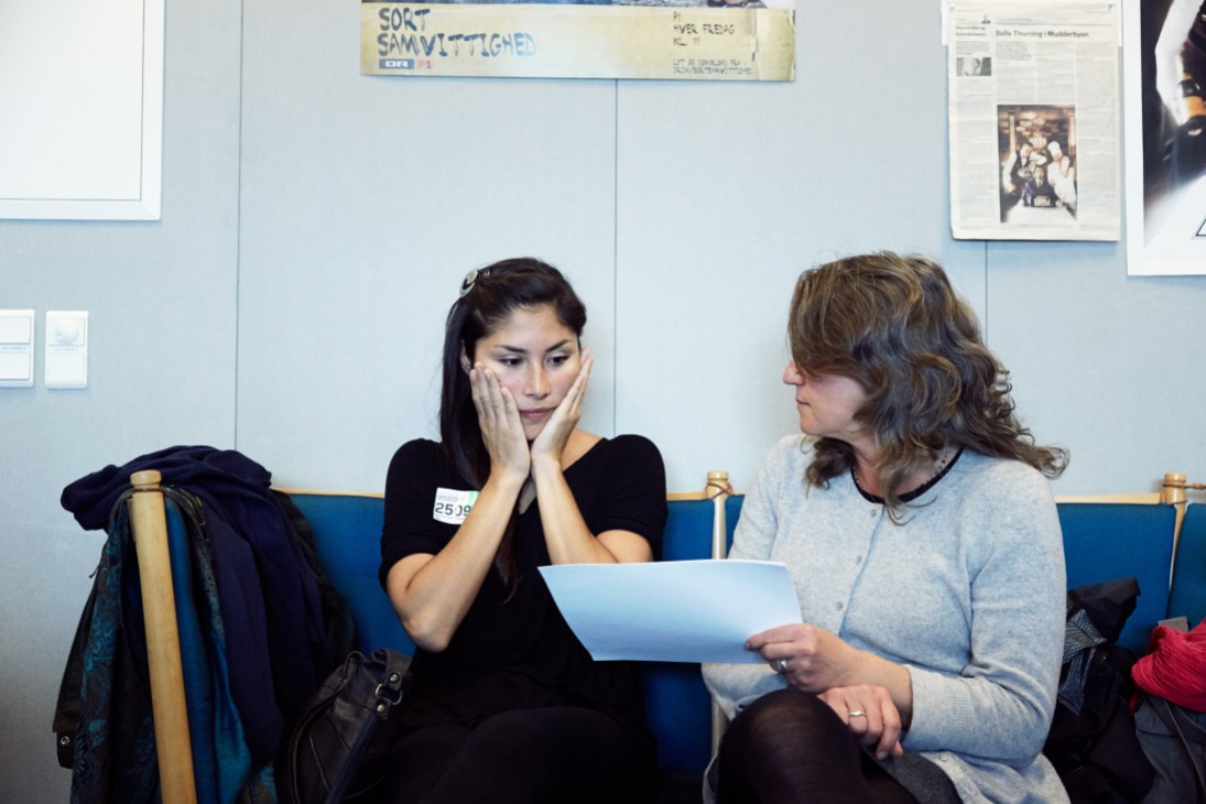 Two women sitting on a blue bench reviewing documents in an institutional setting.