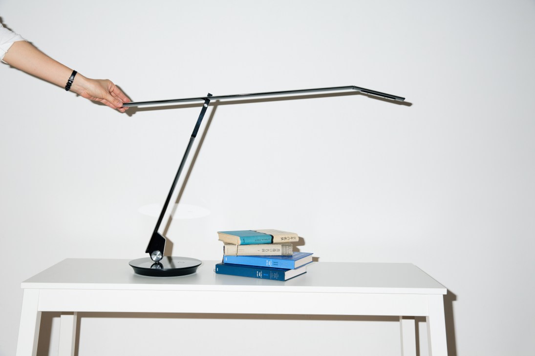 Hand adjusting Balmuda's Airline LED lamp on a desk next to a stack of books.