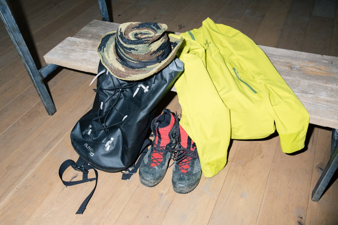 Mountain climbing gear including a straw hat, black backpack, yellow jacket, and hiking boots arranged on wooden floor.