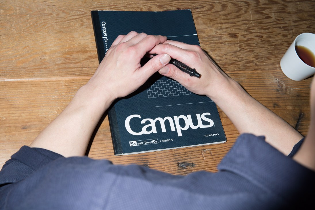 Hands holding a black Campus notebook and pen on a wooden desk with a cup of tea.
